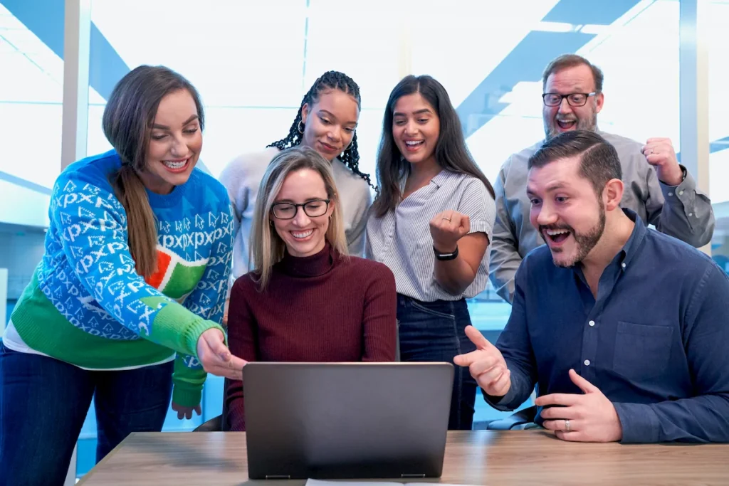 A diverse group of smiling professionals gathered around a laptop in an office, celebrating teamwork and success.
